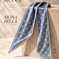 A MONA Bella silk scarf in a dusty blue-gray color with a repeating cream monogram pattern, draped elegantly over a beige stone pedestal. The image features elegant typography on the left and a vertical row of color swatches on the bottom right.